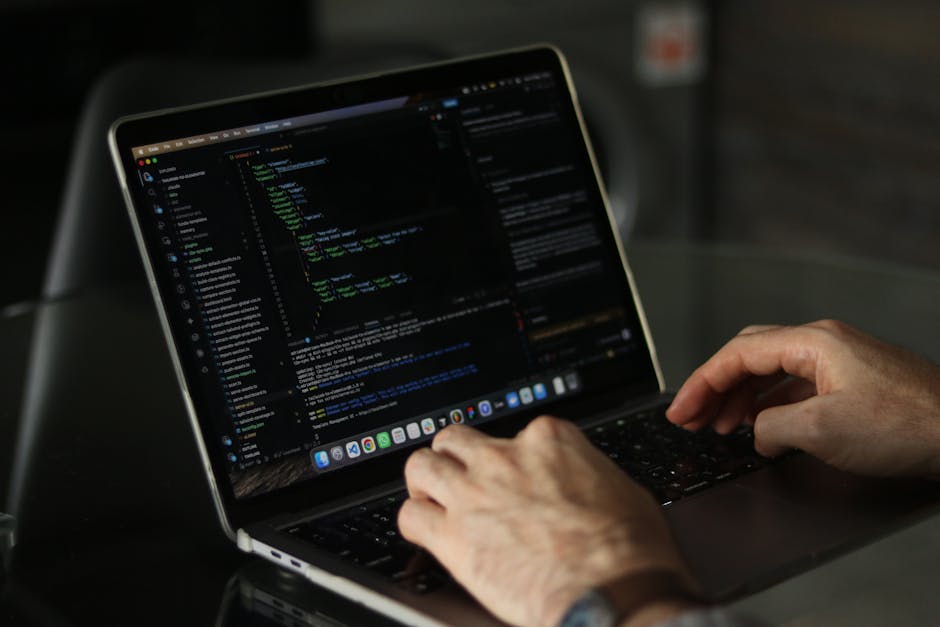 A person working on a laptop with a dark-themed code editor on the screen, showing lines of programming code highlighted in green, blue, and white text. The laptop has a sleek metallic finish, and the